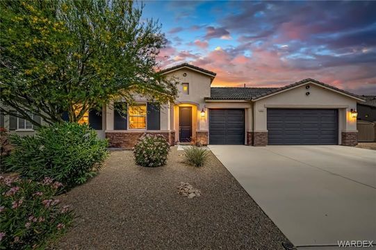 Suburban stucco house at dusk with lit windows, four-car garage, desert landscaping, and pink sky
