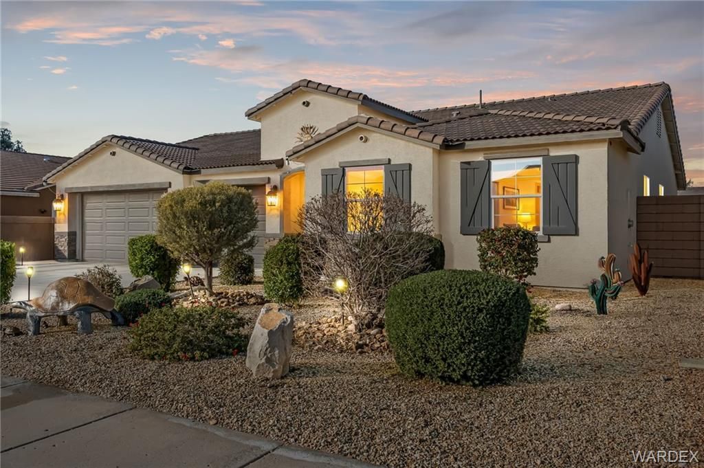 Single-story stucco house at dusk with lit windows, desert landscaping, and a two-car garage