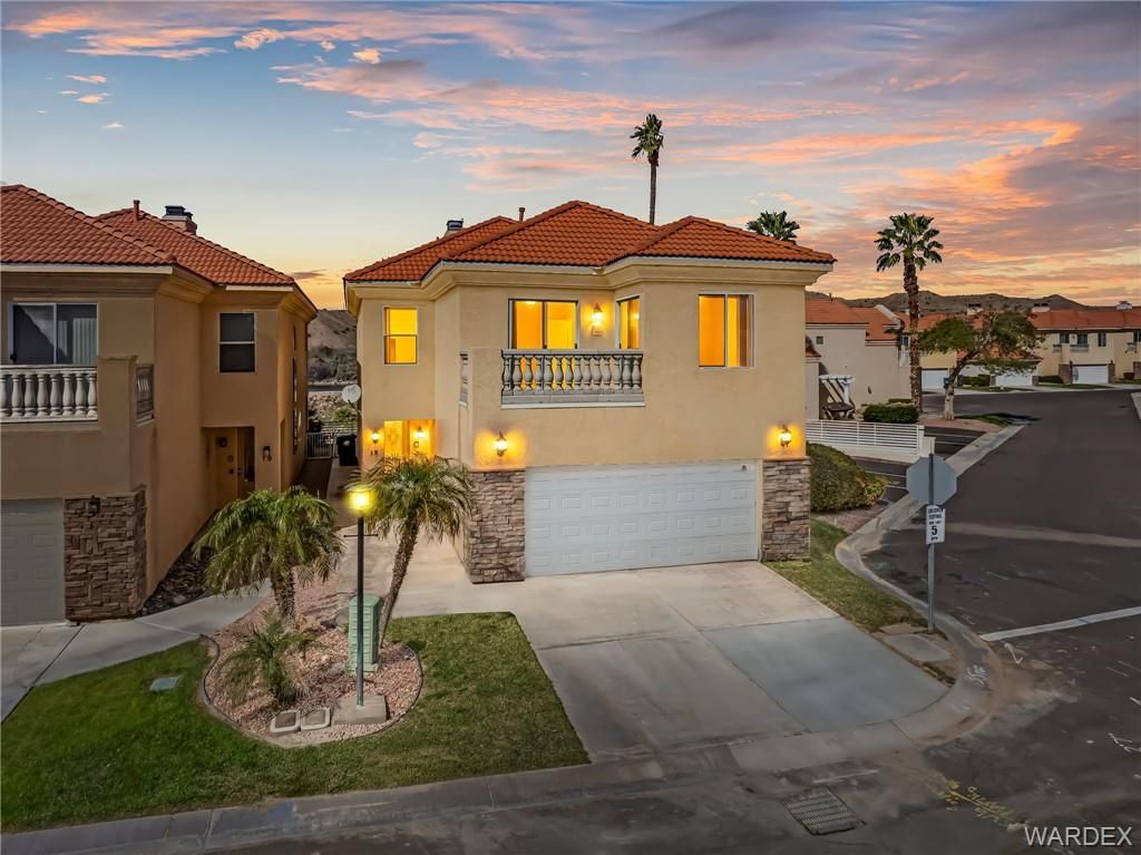 Two-story beige house with red tile roof at dusk, lit windows, and a driveway in a suburban neighborhood
