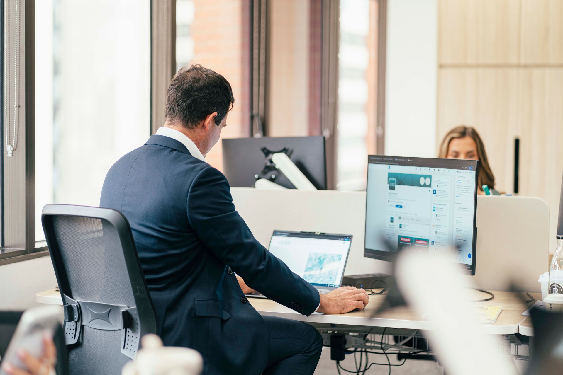 Office worker typing at a desk with dual monitors while a colleague works in the background