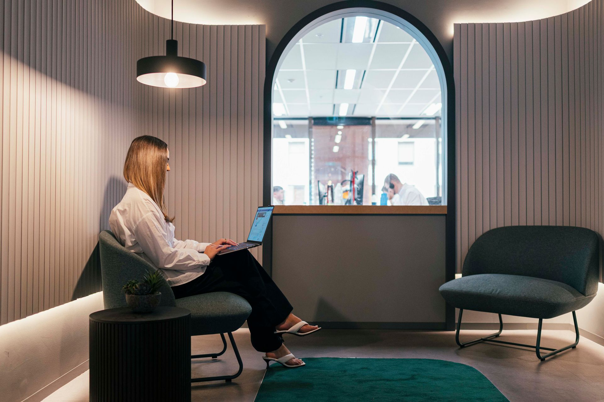 Person sitting in a modern lounge using a laptop, with arched window and chairs nearby