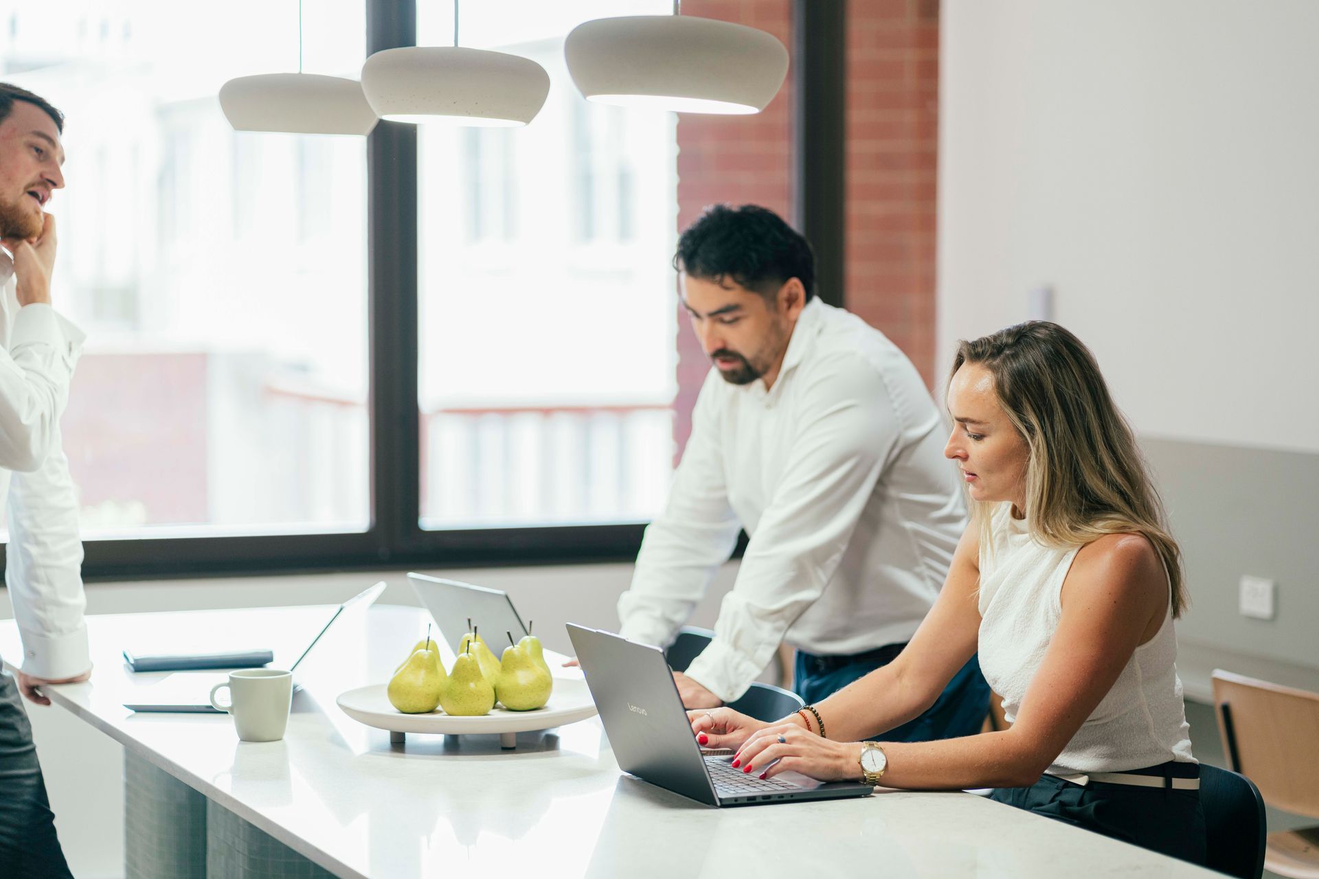 Two coworkers collaborating at a laptop in a bright office, with a bowl of pears on the table