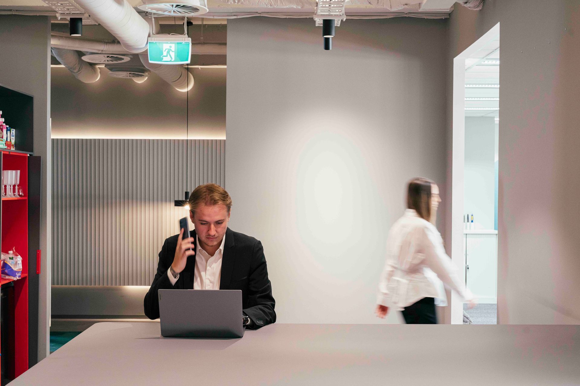 Person working on a laptop in a modern office, while another person walks past in the background.