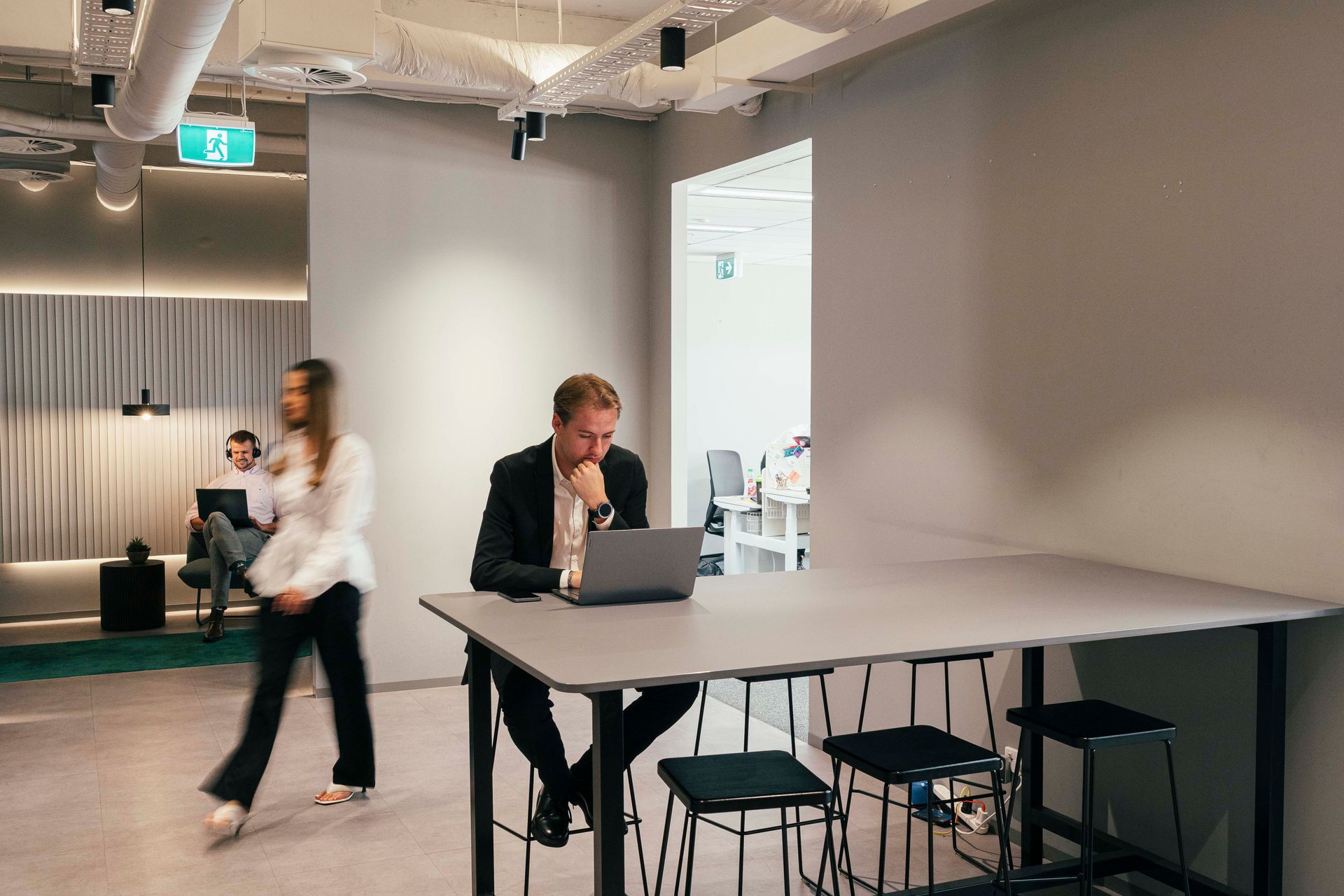 Open office with a person working at a laptop while another walks by near a large table
