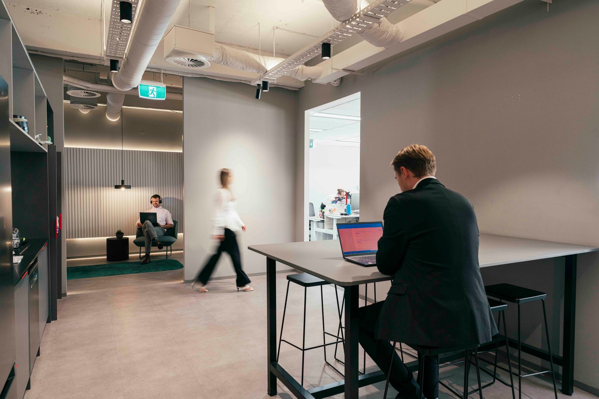 Modern office meeting room with a person working on a laptop at a high table and others in the background