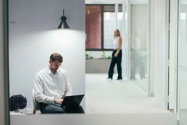 Man using a laptop in a bright office hallway, with a blurred person in the background