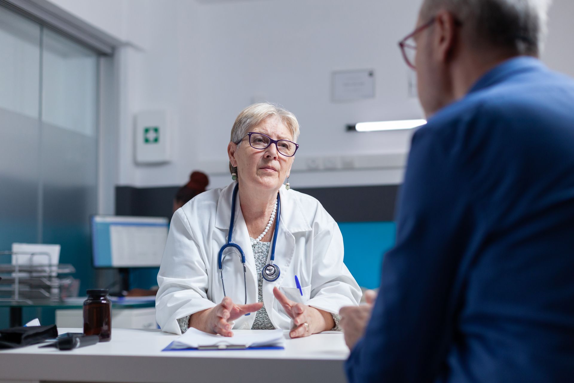 Doctor in white coat speaking across a desk to a seated patient in a bright clinic office.