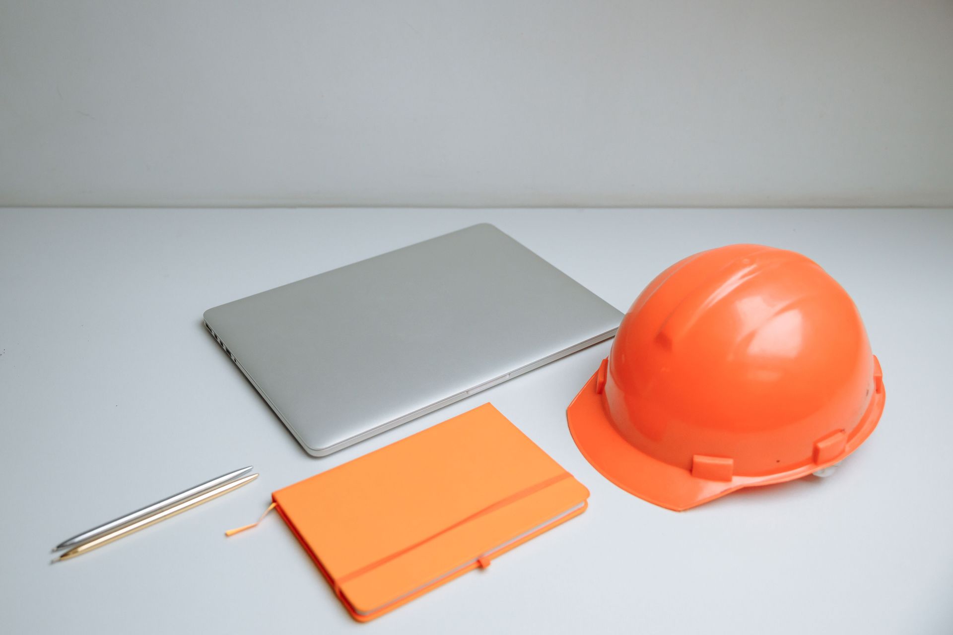 Orange hard hat, gray laptop, orange notebook, and pencil on a white desk
