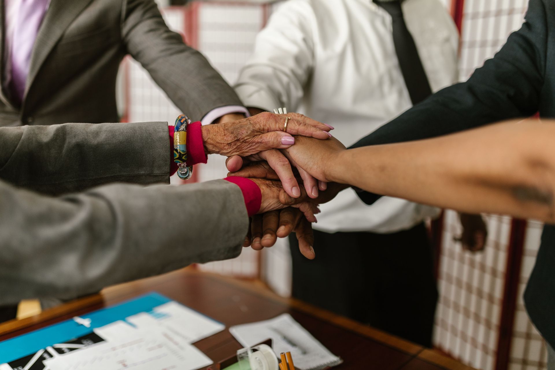 Several hands stacked together over a table in a teamwork gesture, with papers and notebooks nearby.