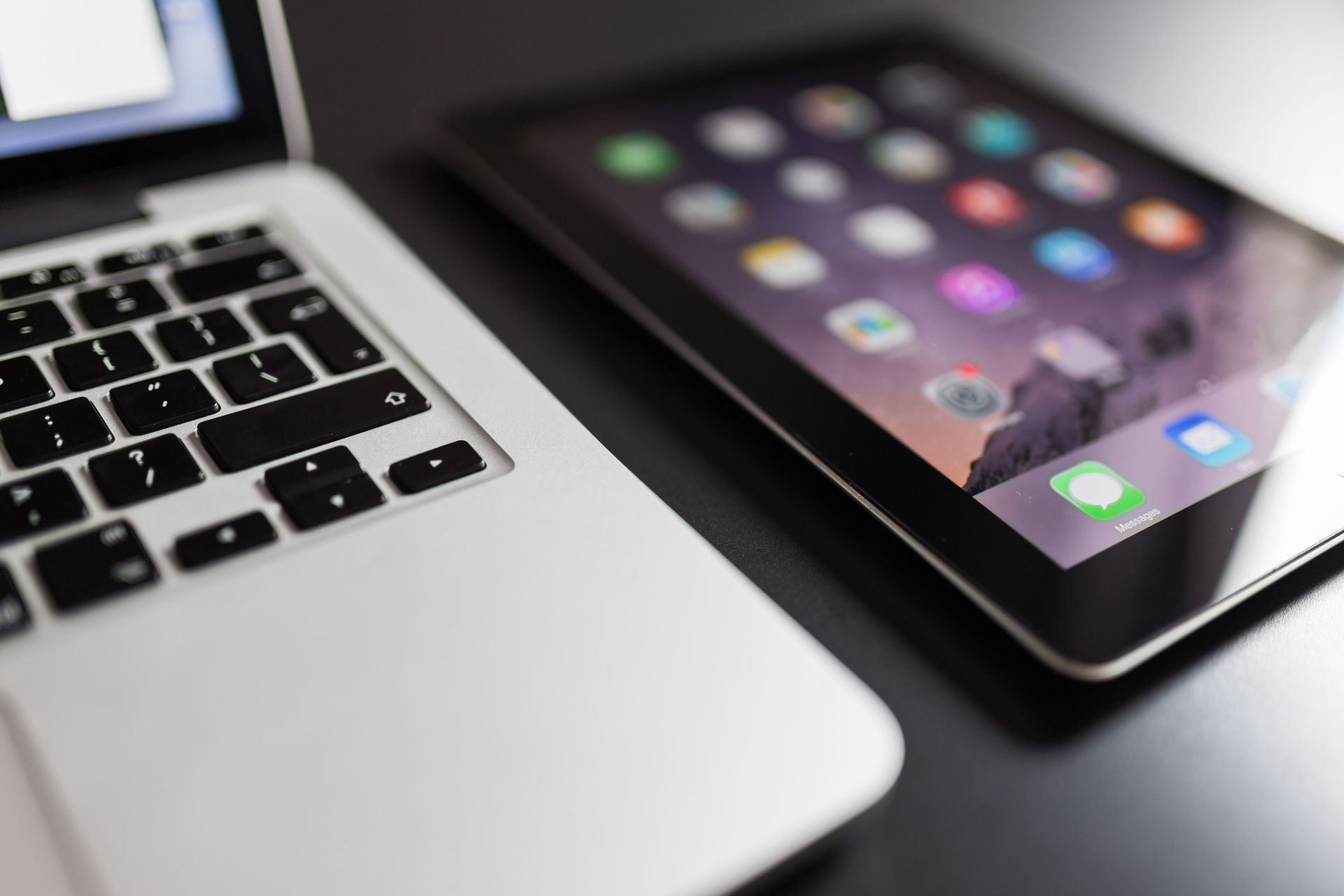 Laptop keyboard beside a tablet showing app icons on a dark desk