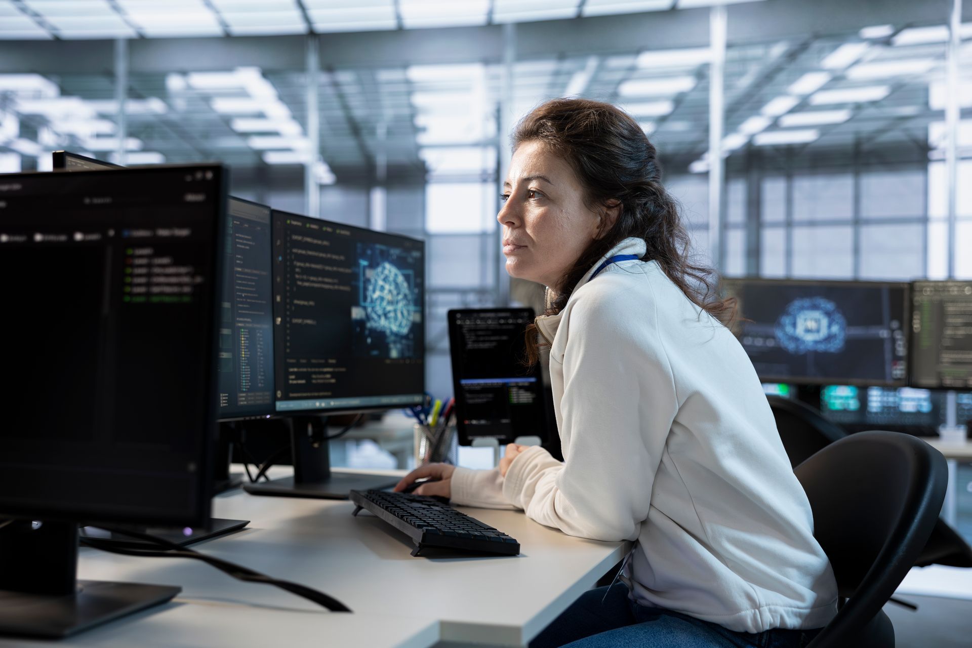 Woman working at a desk with multiple monitors in a bright office, focused on code on screen