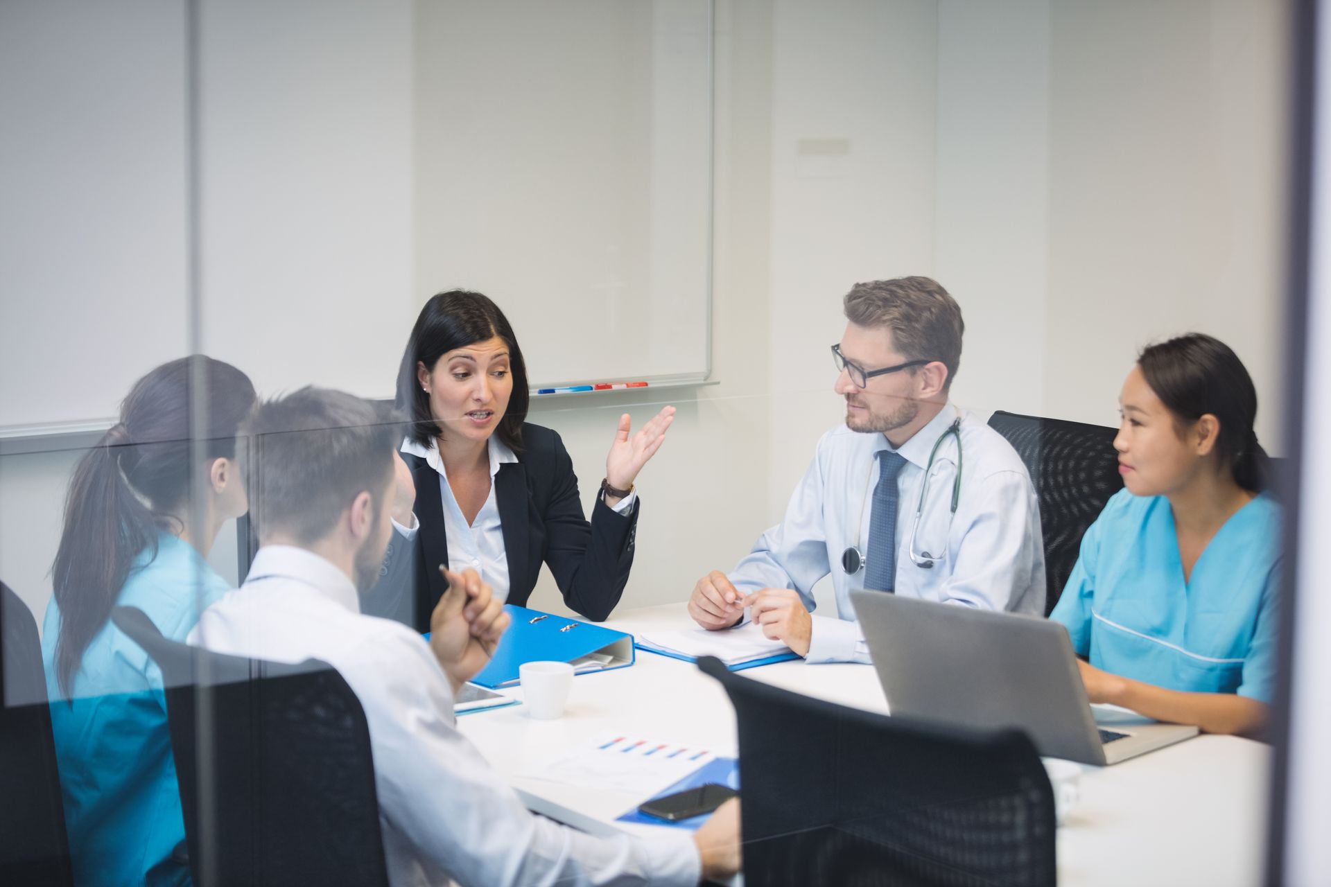 Business meeting in a glass conference room with four people discussing around a table and laptop.