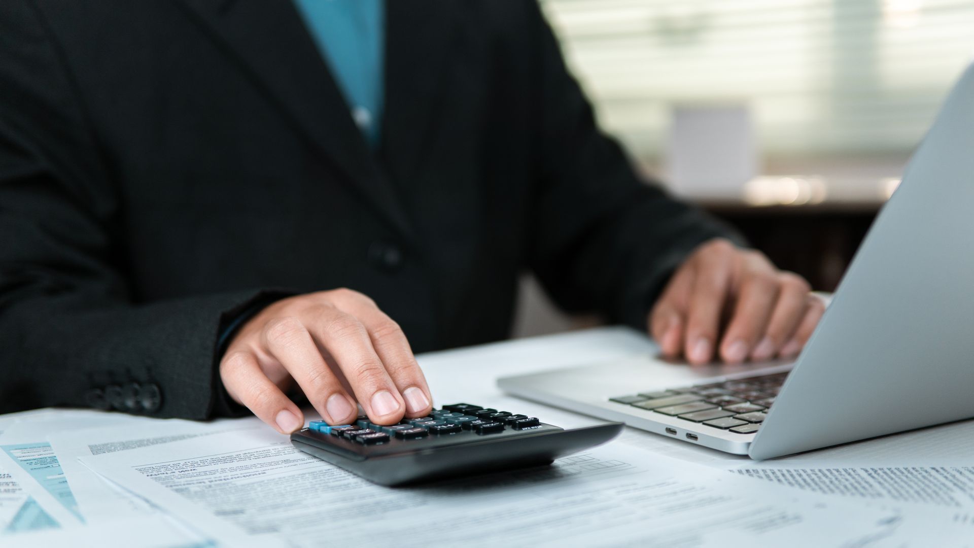 Accountant using a calculator and laptop with forms for business tax preparation.