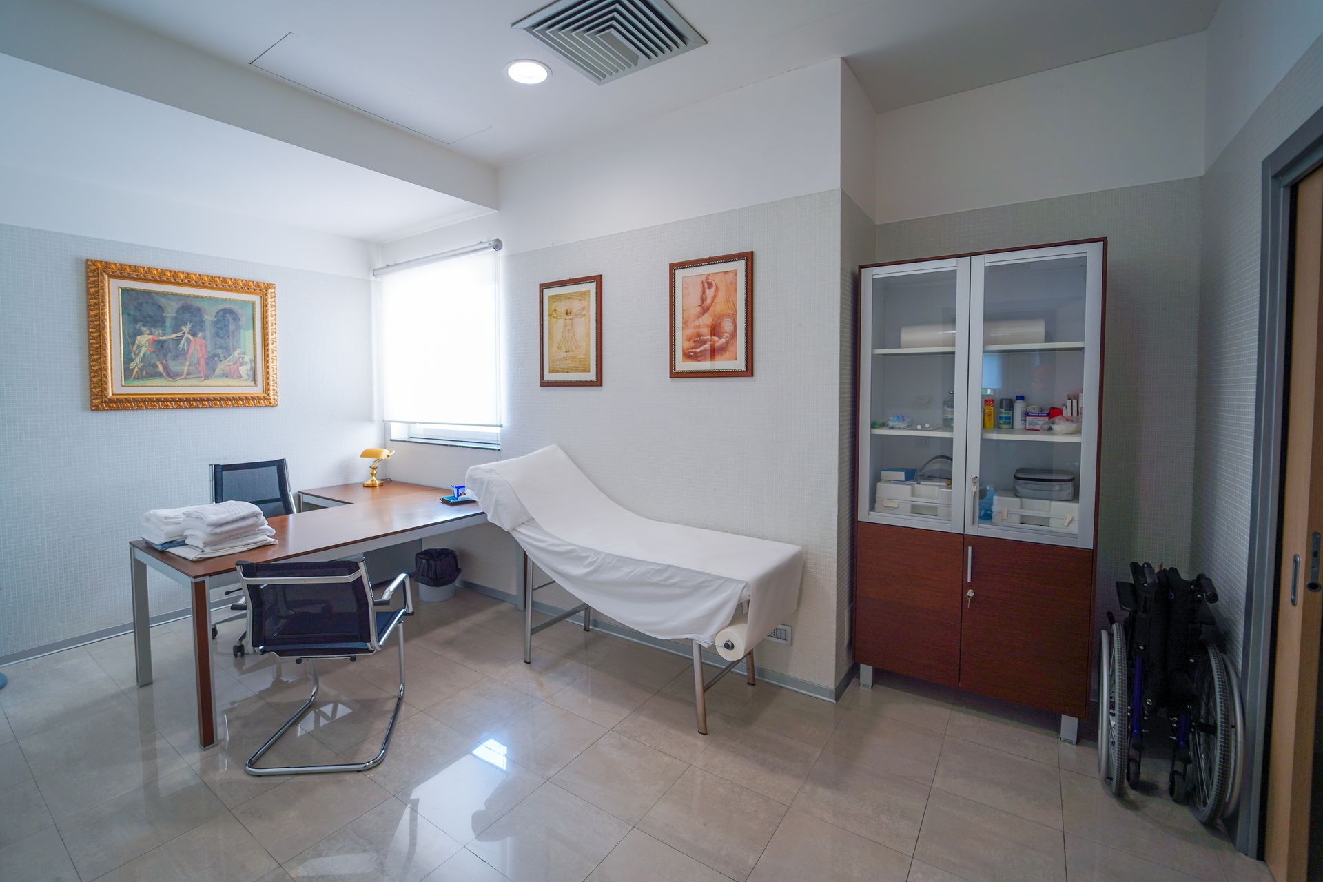 Sala visite mediche con lettino, scrivania, sedie e armadietti in un luminoso ambiente bianco.