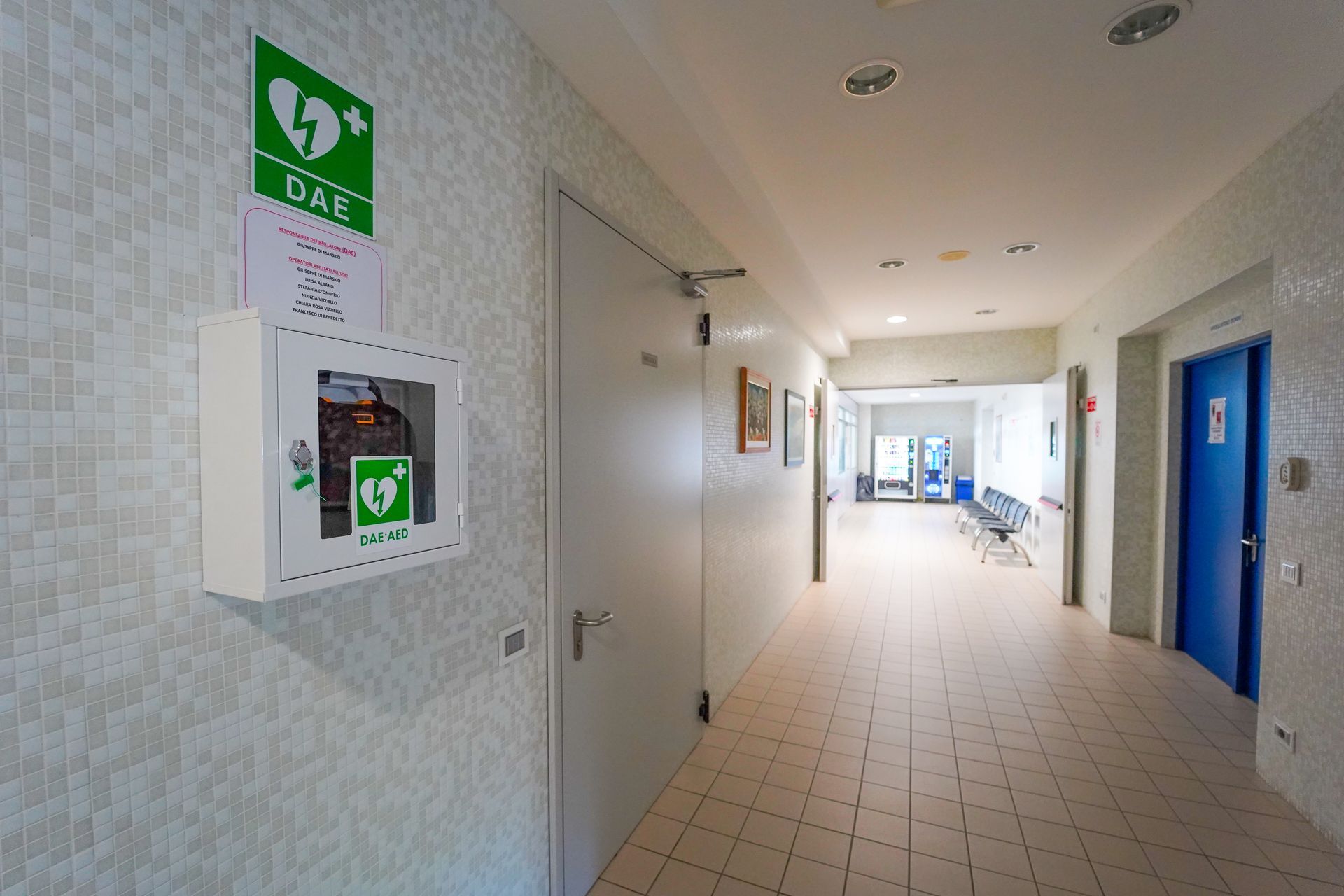 Corridoio luminoso con cartelli di emergenza verdi, una cassetta di allarme antincendio e porte blu sulla destra.