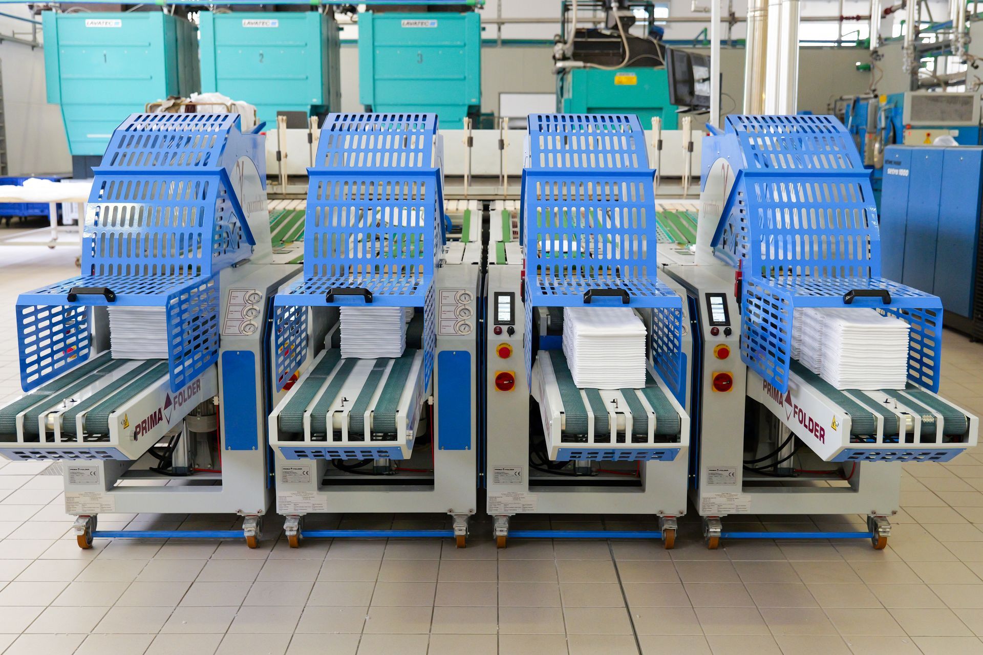 In una fabbrica, quattro macchine industriali blu, ciascuna dotata di nastro trasportatore e tramoggia bianca.