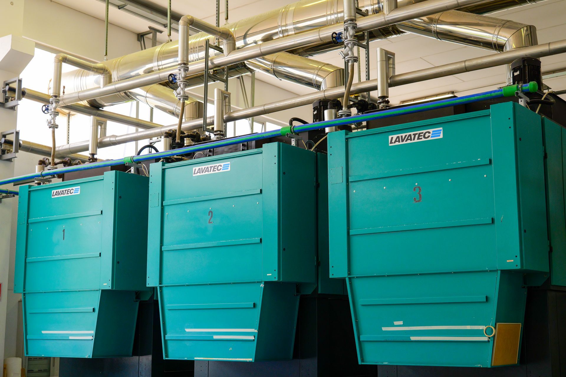 Tre macchinari industriali color verde acqua collegati a condotti aerei in un locale di fabbrica