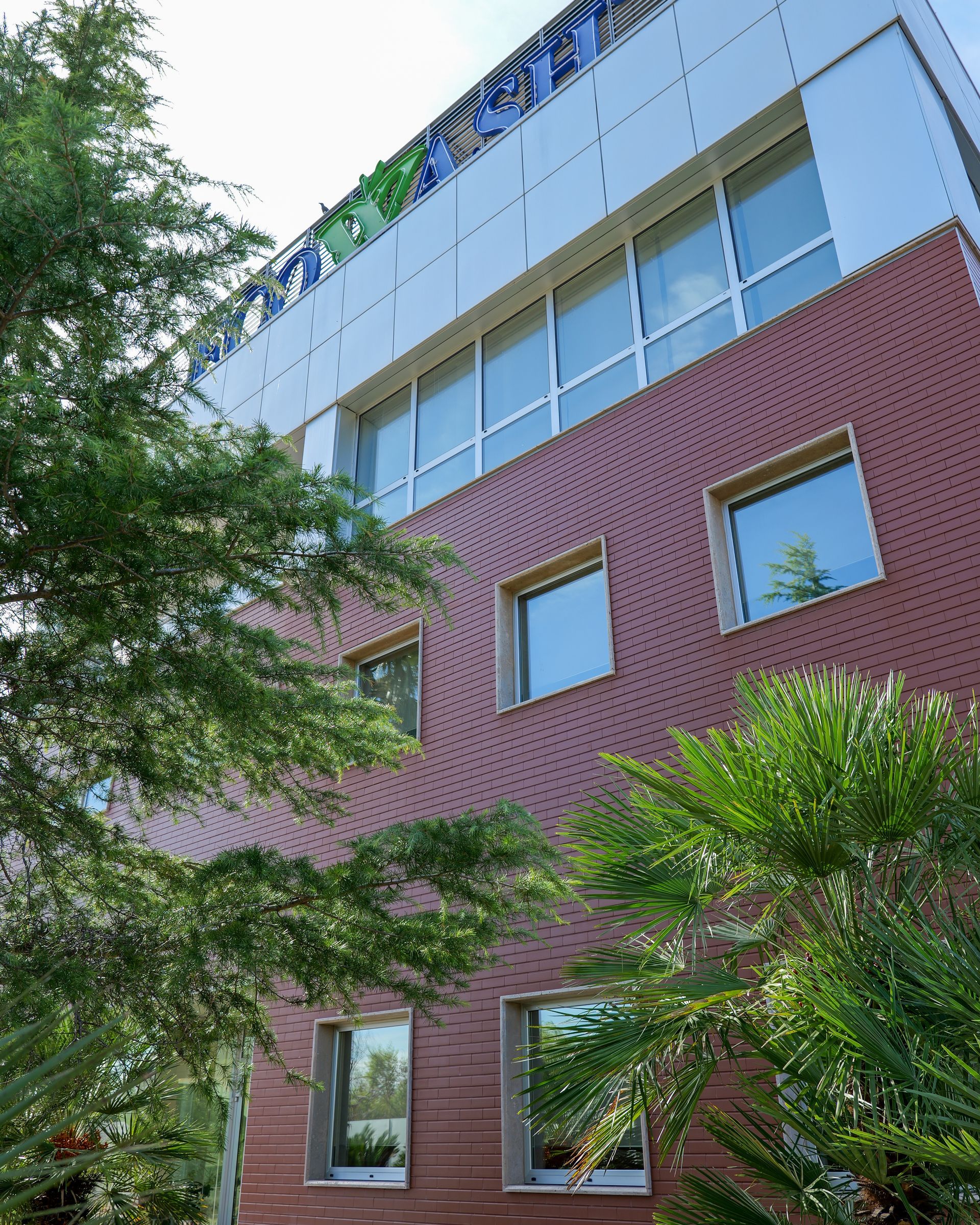 Moderno edificio a più piani con mattoni rossi e vetro blu, parzialmente incorniciato da alberi verdi.