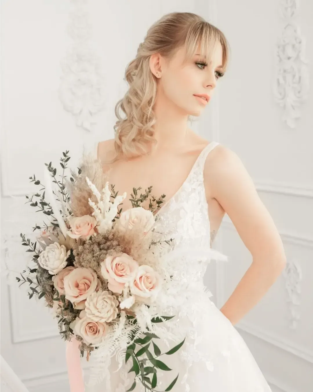 Woman in a white wedding dress holds a bouquet of light pink and cream flowers, posing in a room with white decorative walls.