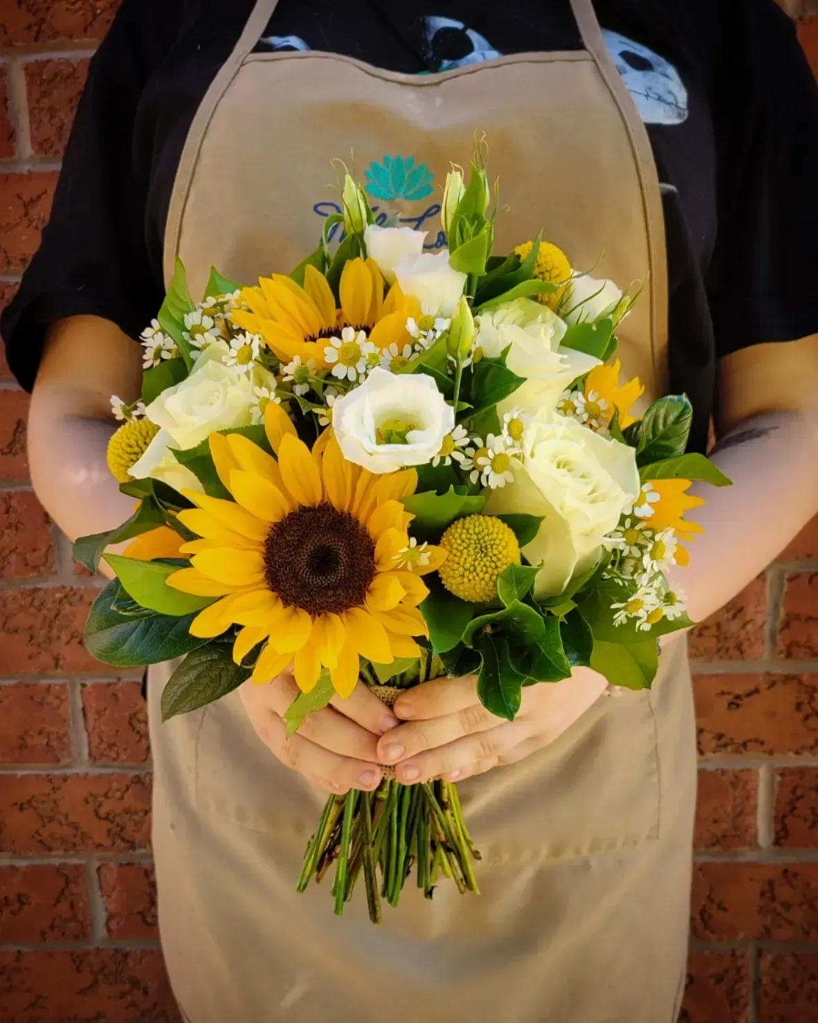 Person holding a bouquet of yellow sunflowers, white roses, and greenery, against a brick wall.