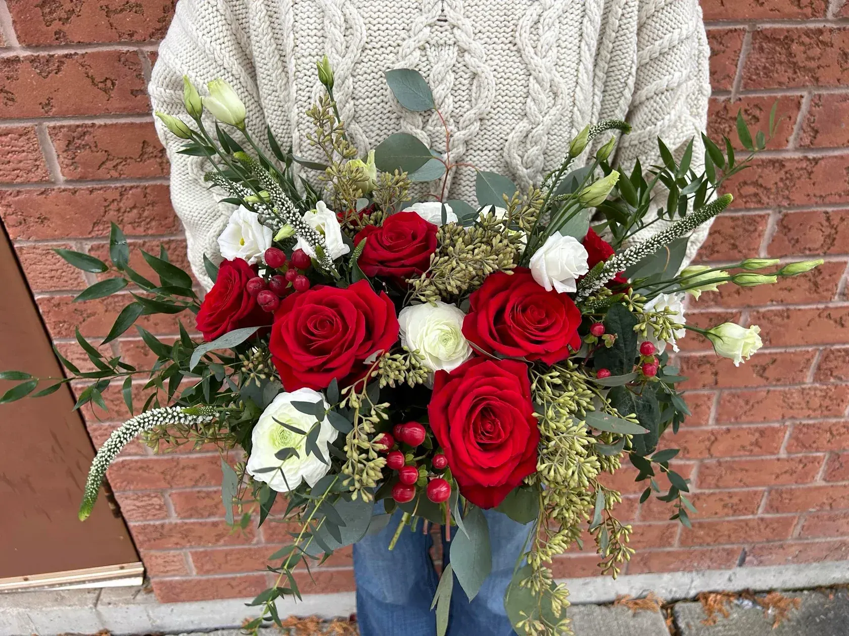 Person holding a bouquet of red and white roses, greenery, against a brick wall.