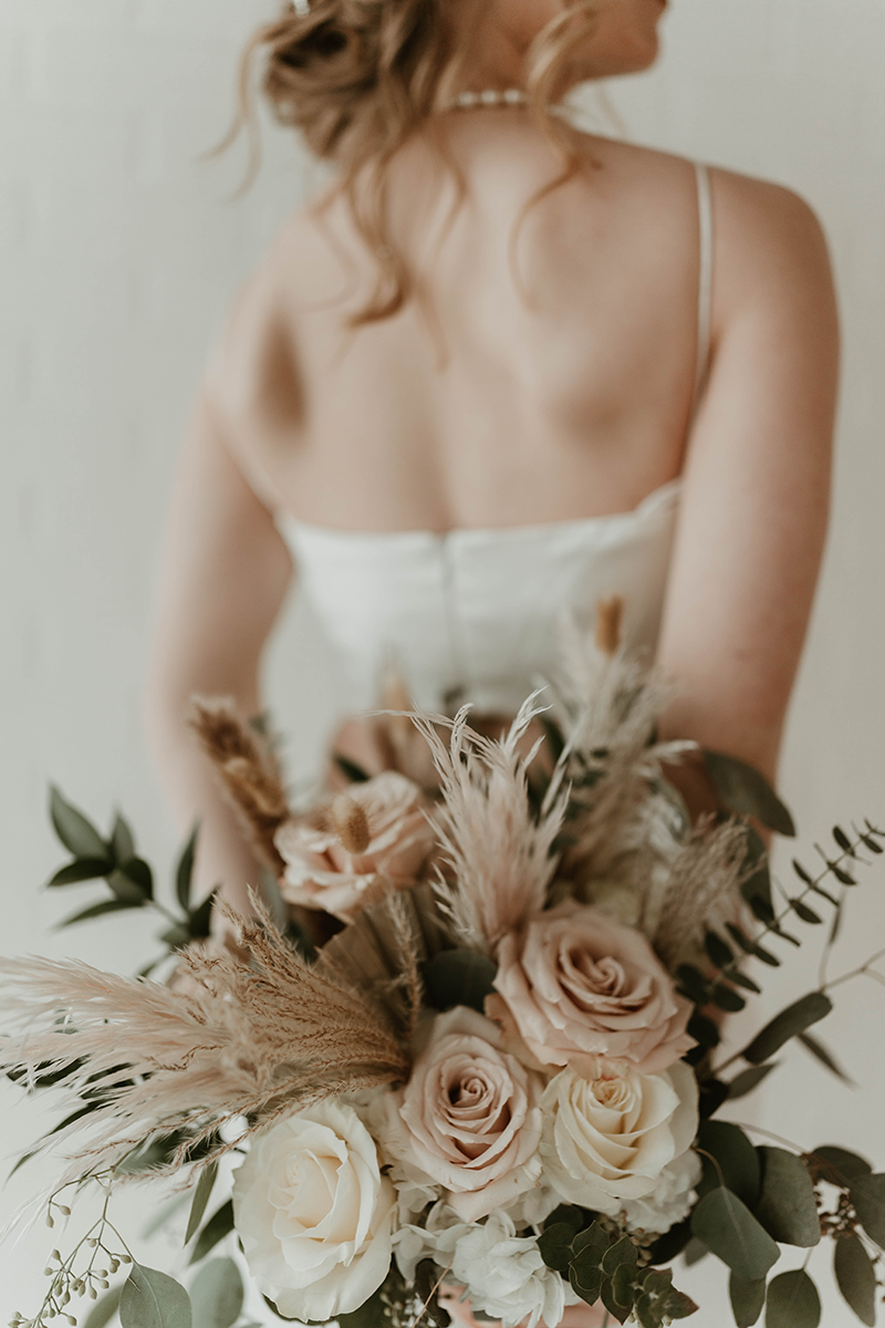 Bride in a white dress holds a bouquet of roses, pampas grass, and greenery; shot from behind.