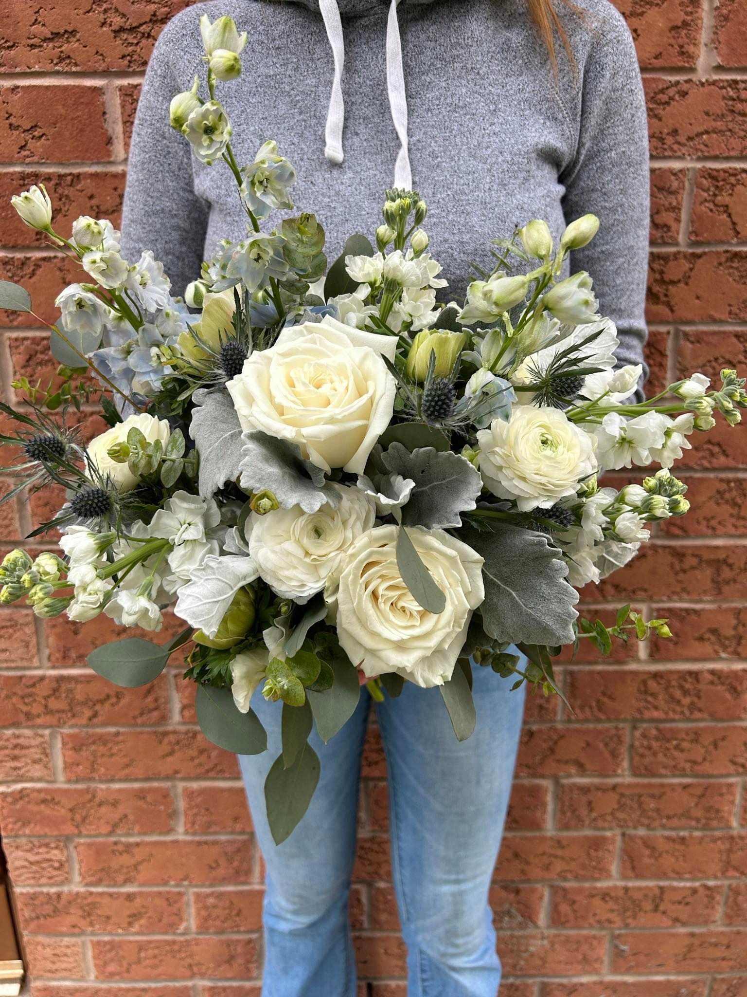 Person holding a large bouquet of white flowers with green foliage, in front of a brick wall.
