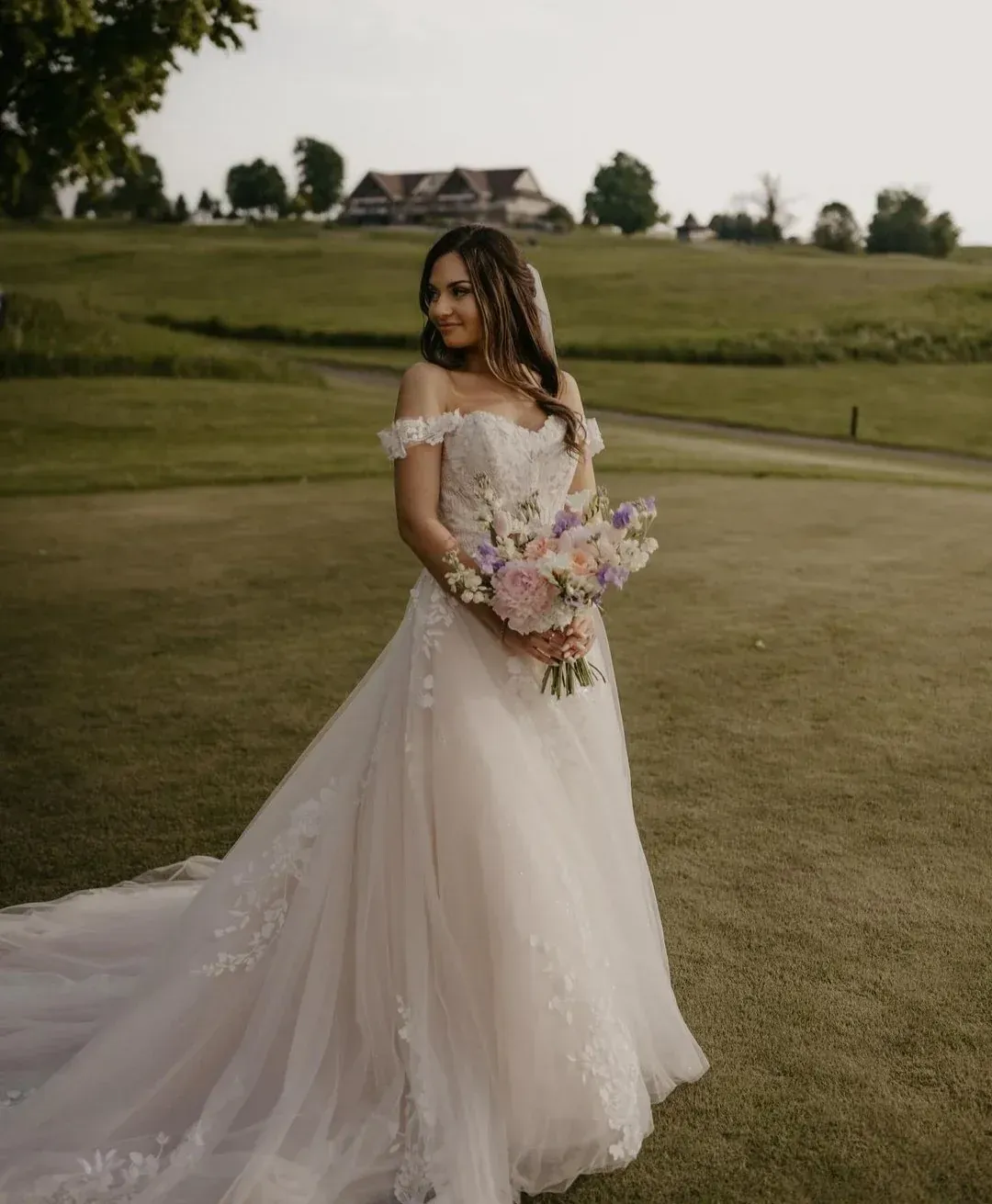 Bride in a white gown holding flowers, posing on a golf course with a clubhouse in the background.