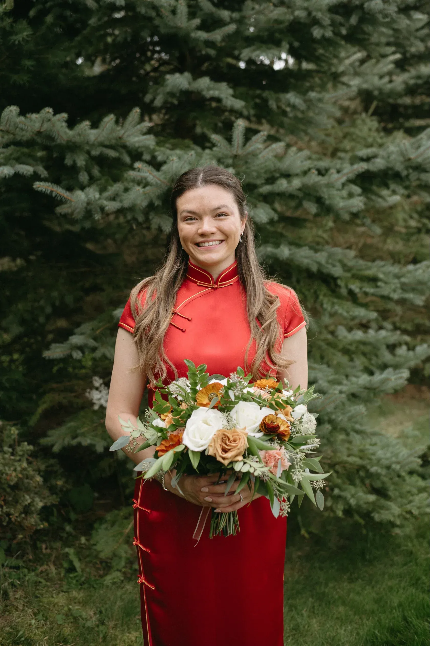 Woman in red cheongsam dress holds bouquet, smiles in front of green tree.