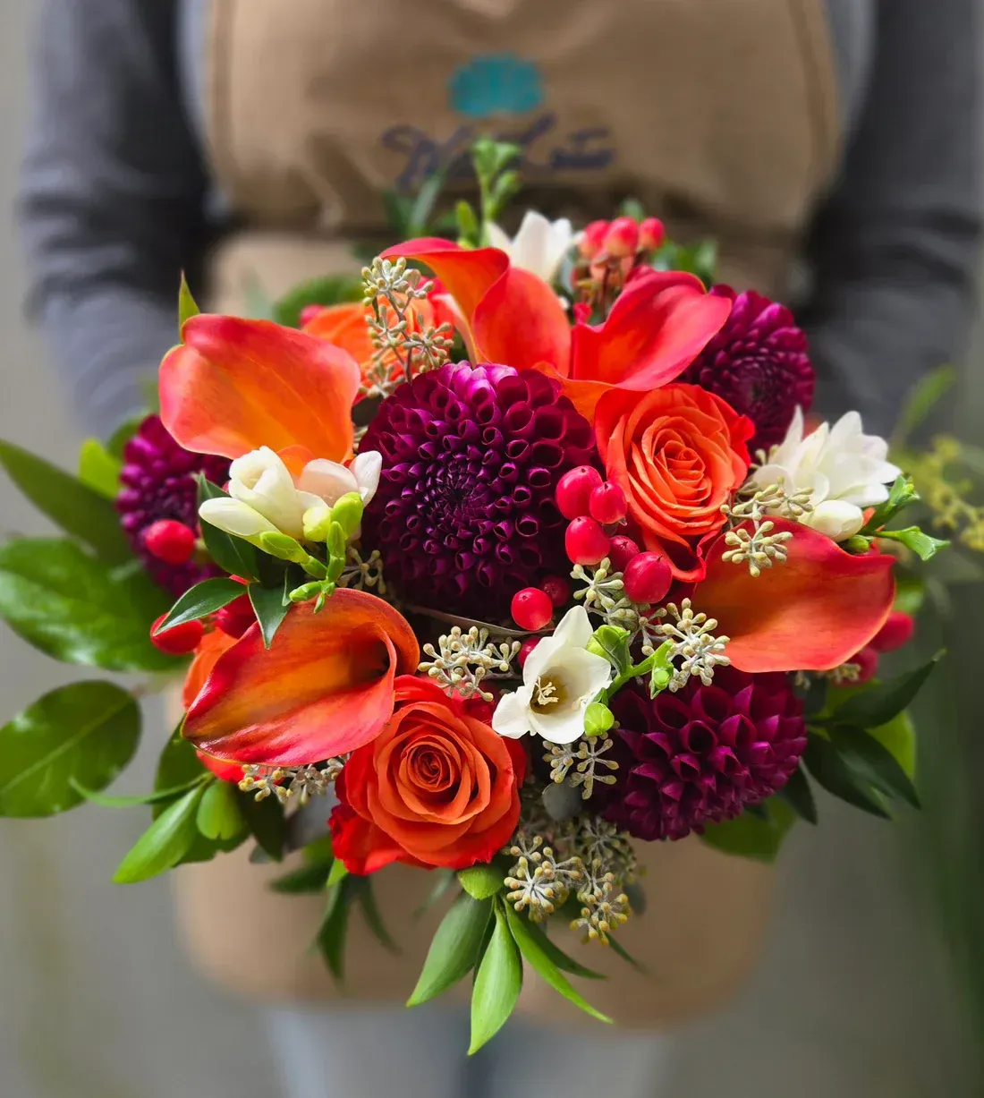 Vibrant bouquet of orange calla lilies, roses, and dahlias held by a person in a beige apron.