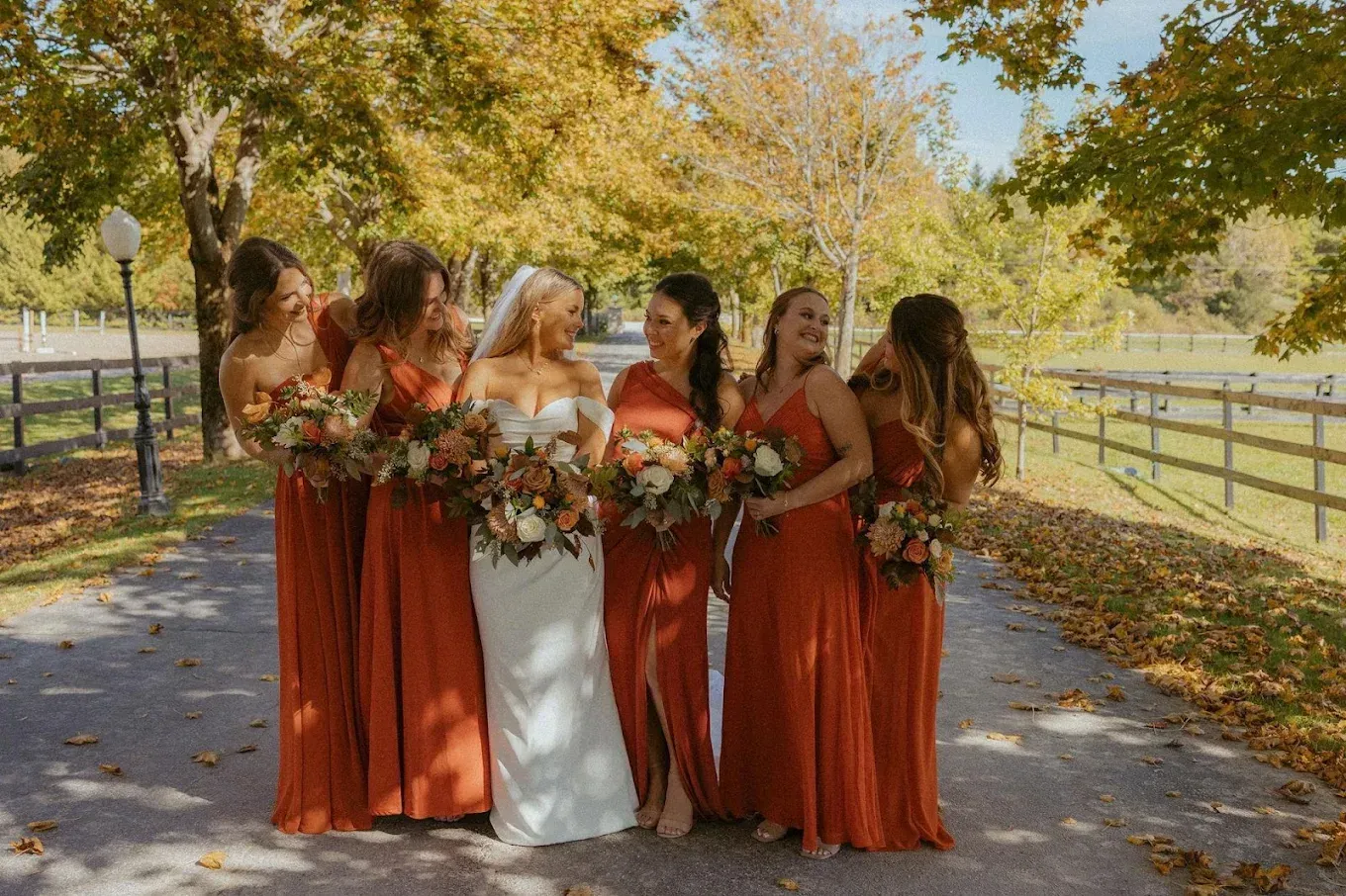 Bride in white gown with bridesmaids in rust-colored dresses, holding bouquets, on a paved path lined with fall foliage.