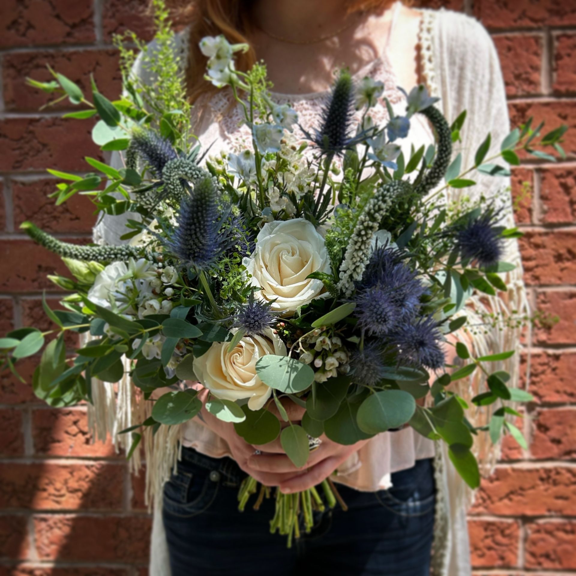 Woman holding a bouquet of white roses, blue thistles, and greenery against a brick wall.
