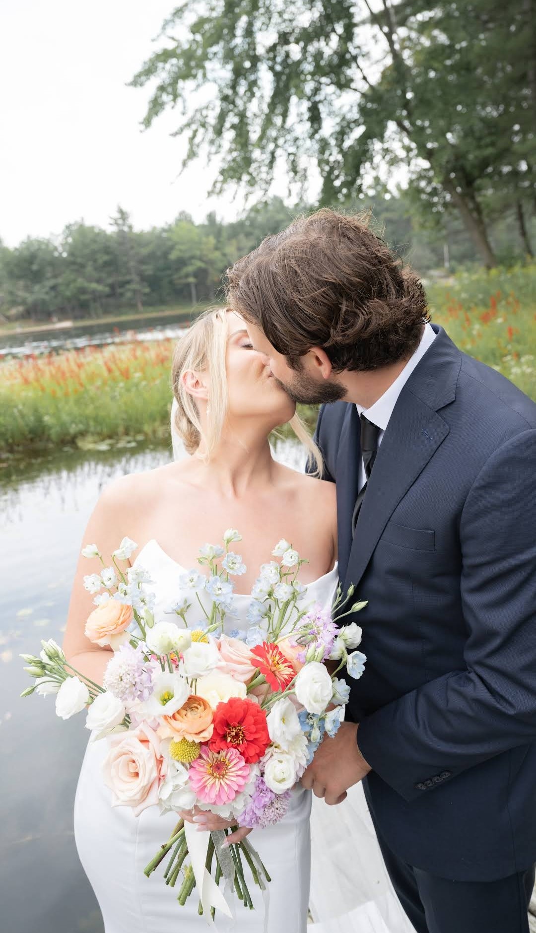 Newlyweds kiss on a dock, holding a colorful bouquet. The bride wears a white gown; the groom, a navy suit.