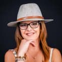 Woman with glasses and a fedora hat smiling, resting her chin on her hand. She has long, red hair. Black background.