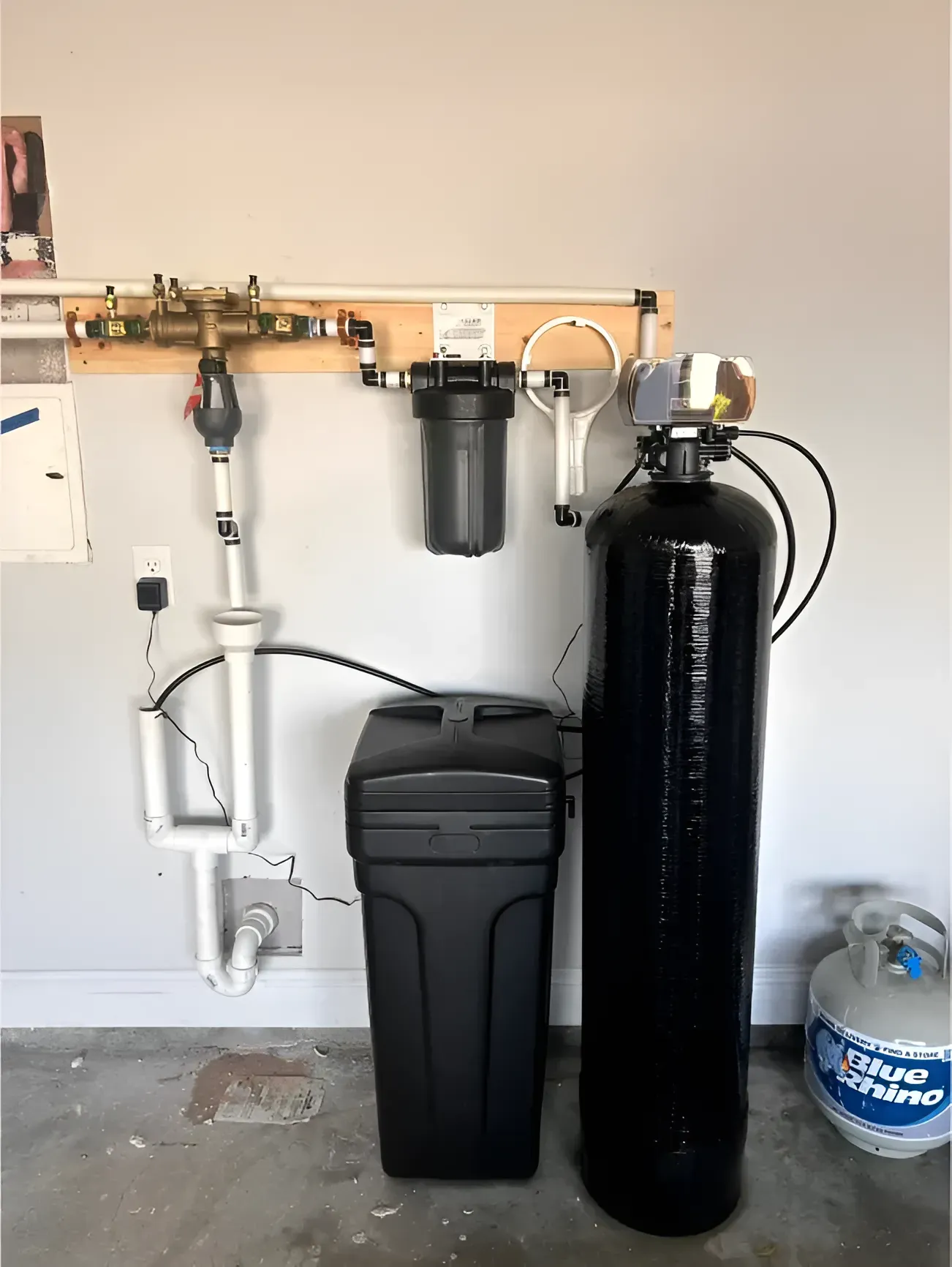 Water softener system installed in a garage; includes a tank, filtration, and salt storage.