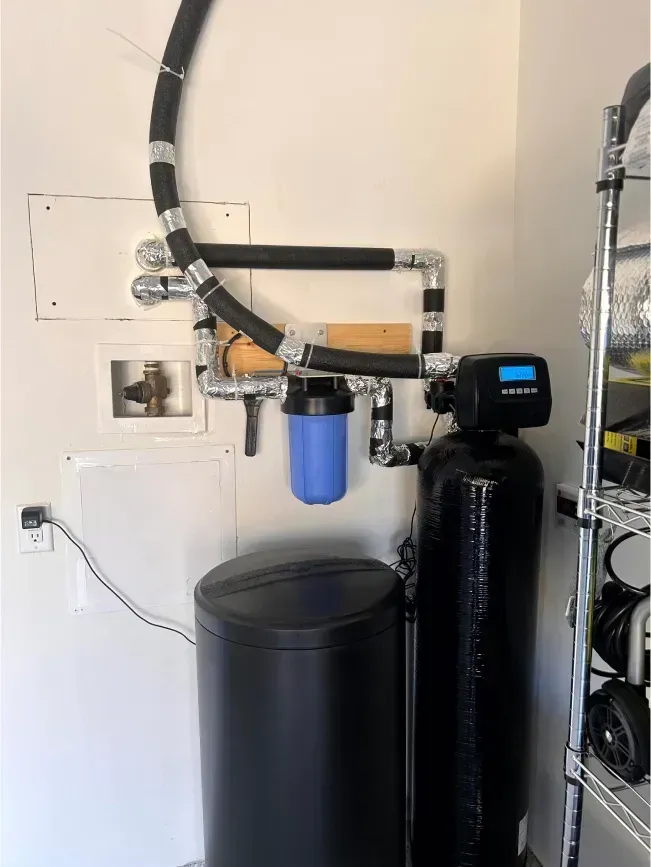 Water softener system in a garage setting with blue filter, black tanks, and silver pipes.