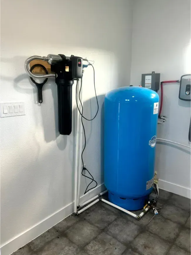 Water filtration system and blue pressure tank installed in a white-walled room, connected by pipes and wires.