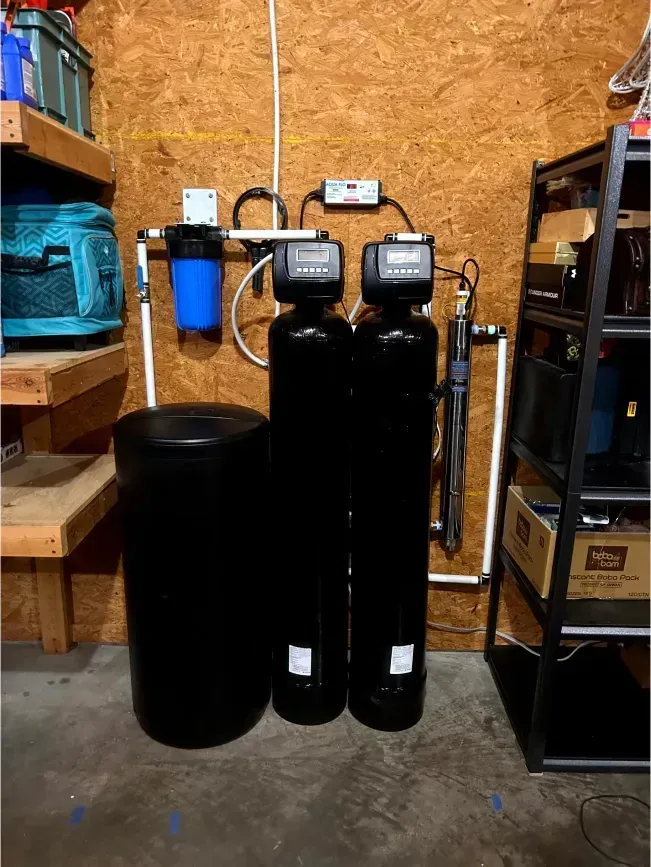 Water filtration system in a garage. Includes a blue filter, black tanks, and a black barrel.