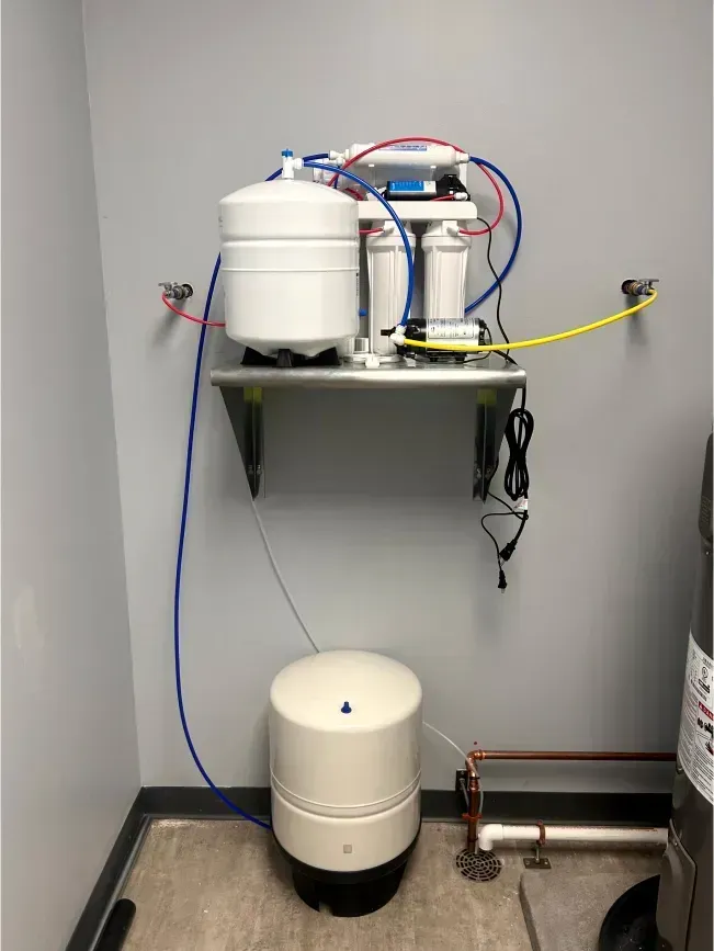 Water filtration system mounted on a wall shelf. White tanks and blue/red tubing. Ground-mounted water storage tank below.