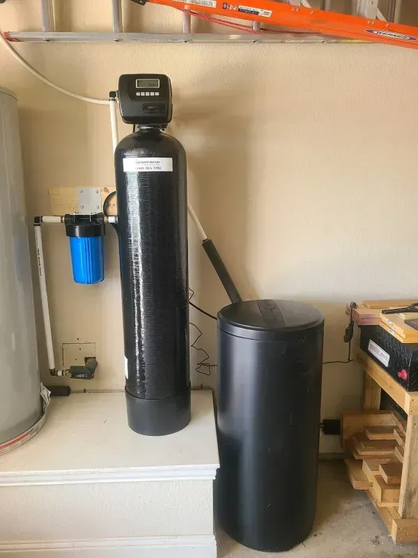 Water softener system in a garage setting with a tall black tank and a blue filter.