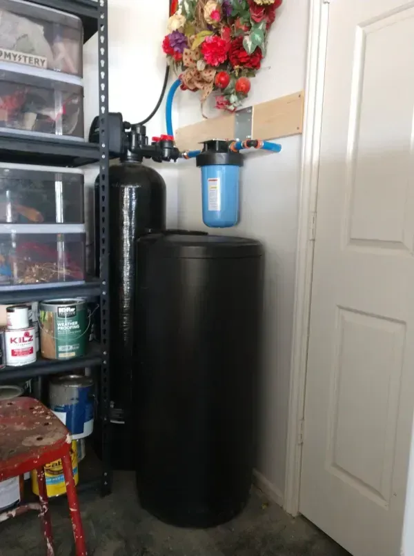 Water softener system in a garage corner; includes a black tank, blue filter, and connected pipes.