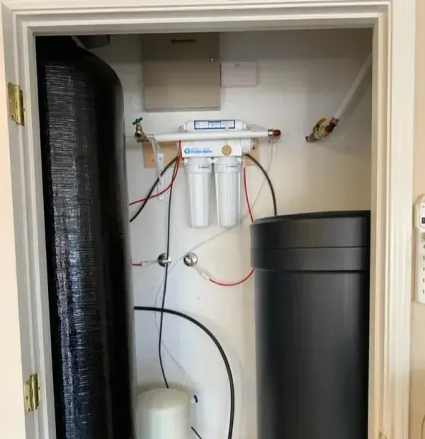 Water filtration system in a utility closet with black tanks, white filters, and plumbing.