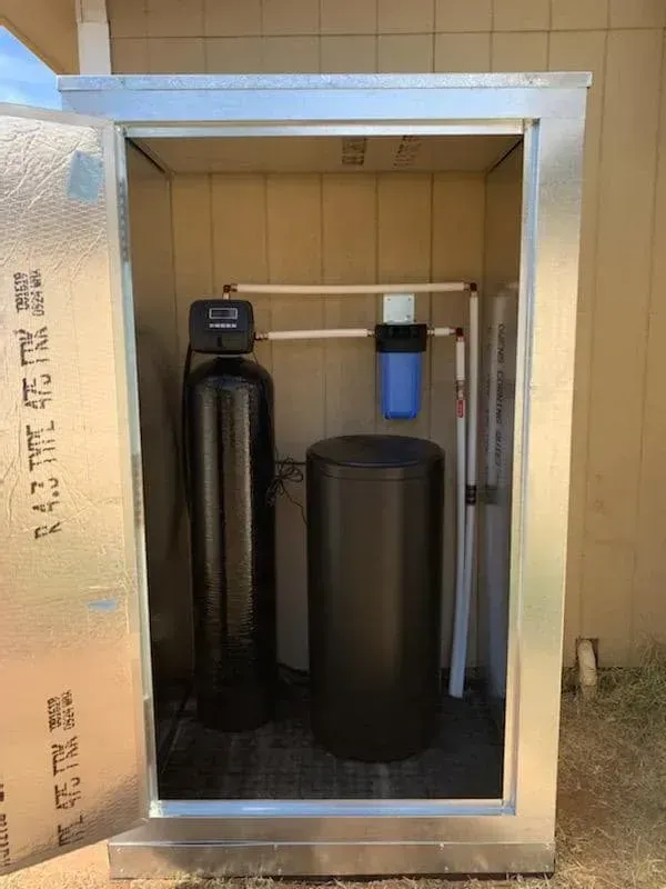 A water filtration system in an outdoor metal cabinet. Features a black water softener tank, filter, and brine tank.