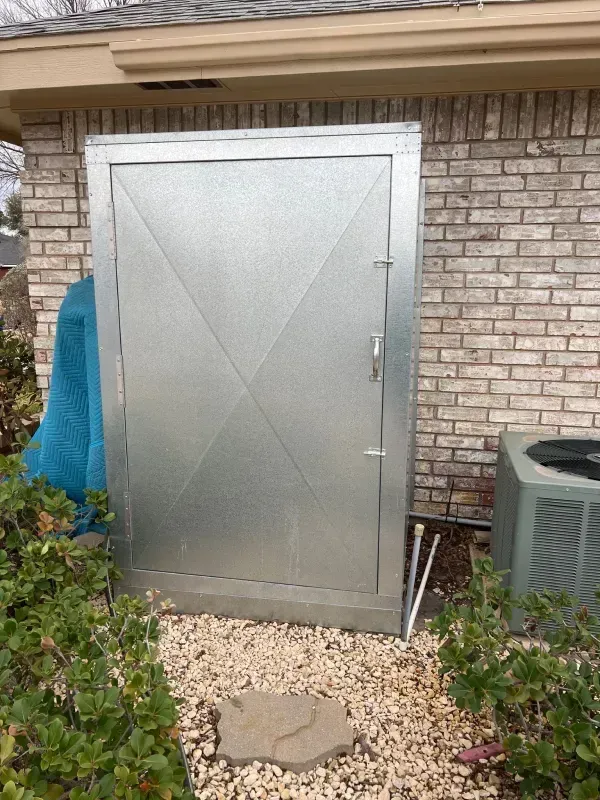 Metal door on brick wall, next to HVAC unit, with gravel walkway and greenery.