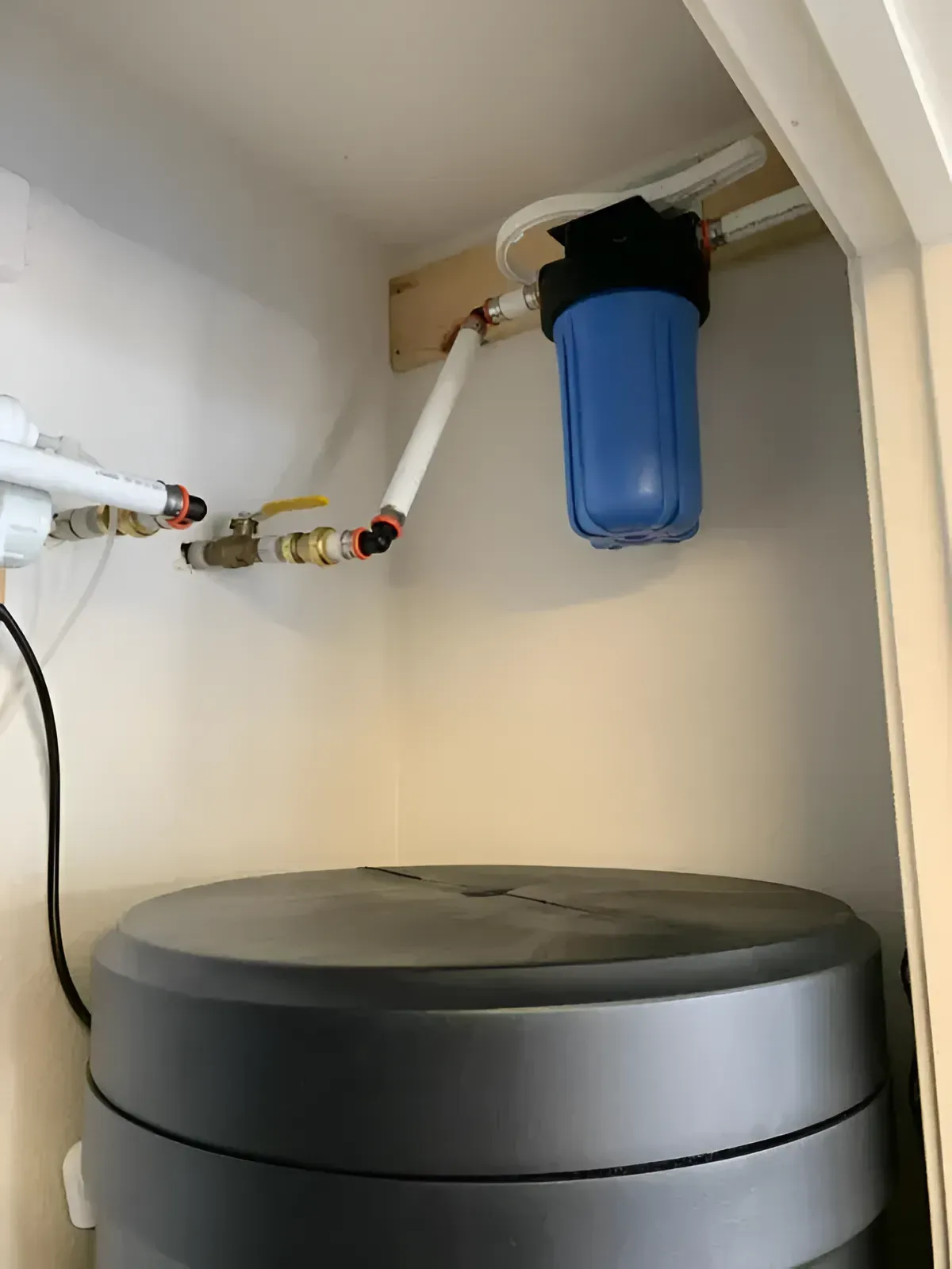 Blue water filter and plumbing mounted above a gray water heater in a utility closet.
