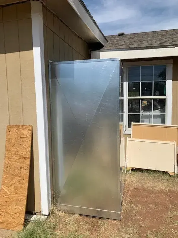Metal air duct leans against a tan building next to a window and wood pieces.