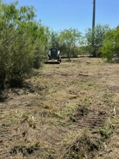 Person mowing a dry grassy field with a small tractor beside trees and utility poles.