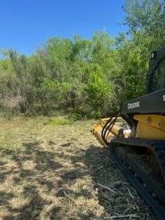 Yellow bulldozer working on dry grassy ground near green shrubs under a blue sky