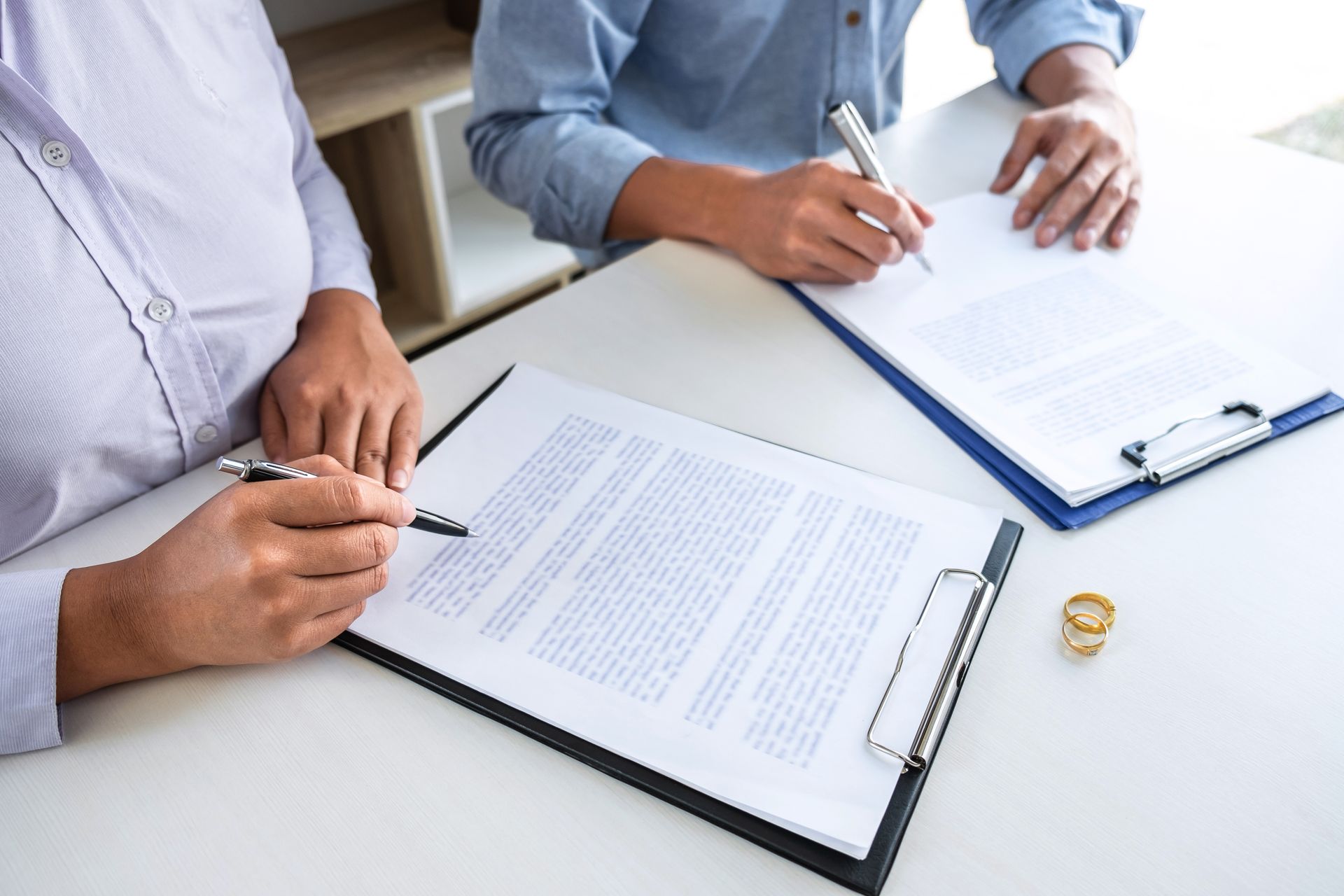 Two people signing legal documents beside a wedding ring set.