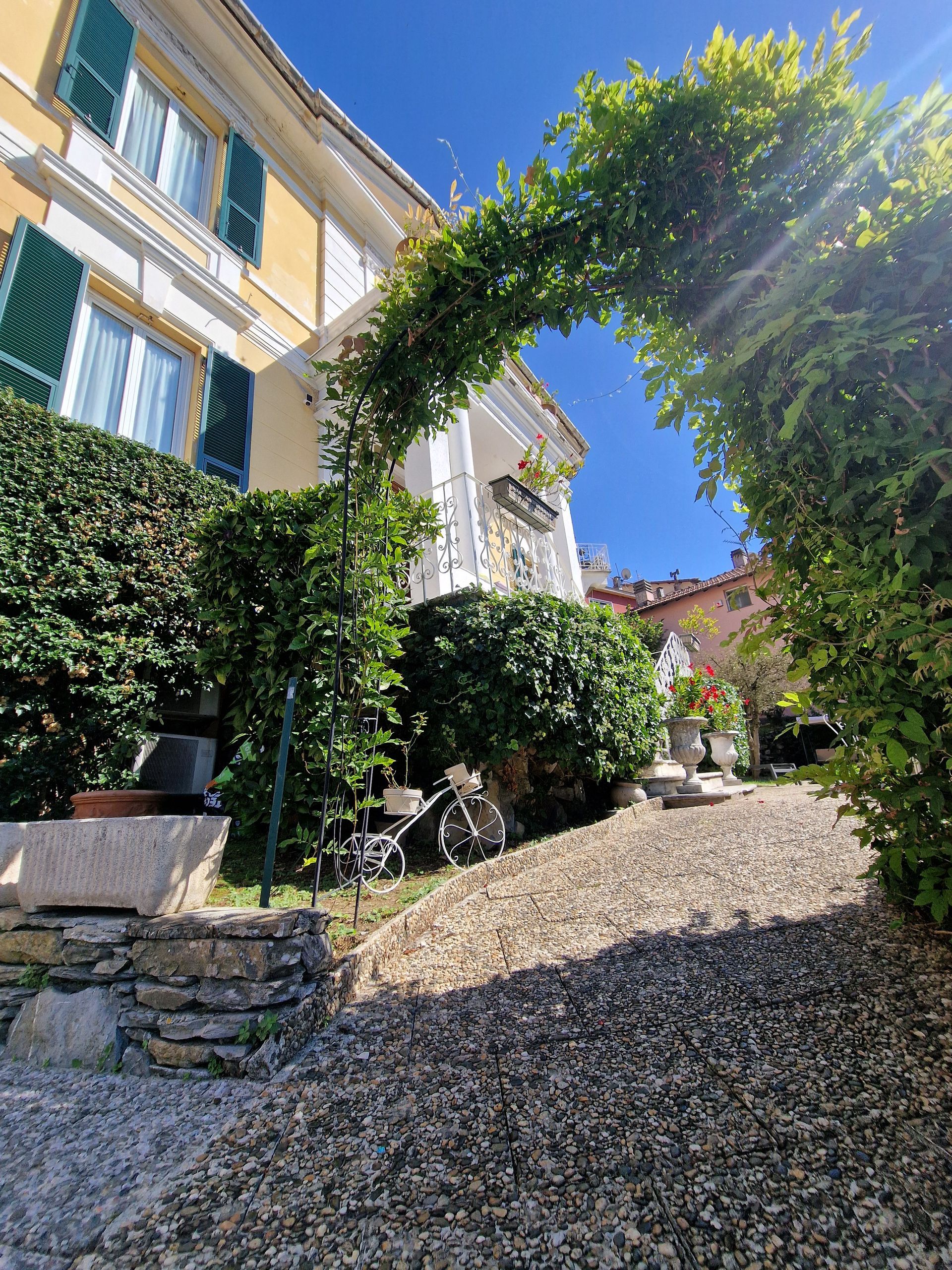 Esterno della casa con sentiero in pietra, arco verde, pareti gialle, persiane verdi, giornata di sole.